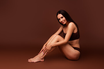 Fototapeta premium Full body photo no retouch of pretty young girl sit floor touch legs spa salon dressed stylish underwear isolated on brown color background