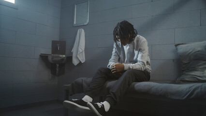 Depressed African American guy sits on the bed in prison cell. Caucasian teenage inmate washes his face in the sink. Young criminals, prisoners serve imprisonment term in jail. Youth detention center.