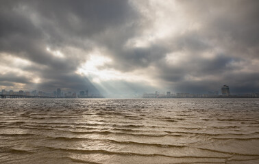 Obraz premium Dnipro River, Dnipro city, Ukraine. Cityscape with sky with clouds and river and city bridge. Cloudy clouds hang over and the sun's rays shine on the cityscape.