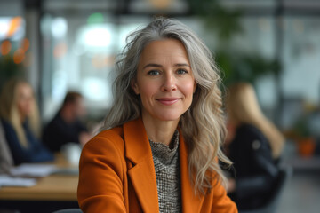 A middle-aged business woman with long gray hair smiles and looks straight. Behind, on a blurred background, business processes take place between office employees. Business woman concept - obrazy, fototapety, plakaty