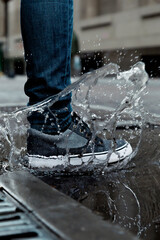 Close-up of a foot with sneakers stomping forcefully on a puddle of water in the street, creating a...