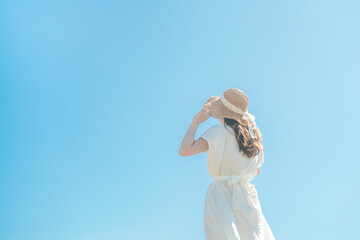 青空の下で遠くを眺める白いワンピースに麦わら帽子の女性