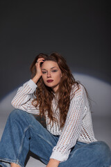 Beautiful fashion youth girl in a stylish white sweater with vintage jeans sits and poses in the studio