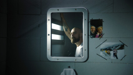 Reflection of male criminal in the mirror looking out the window in prison cell. Pictures of family...