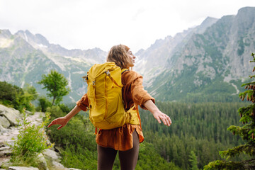 Young woman with a yellow hiking backpack traveling along hiking trails in the mountains among forests and cliffs. Travel, vacation concept.
