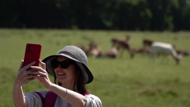 Park with deer. A woman films herself on her phone with deer in the background.