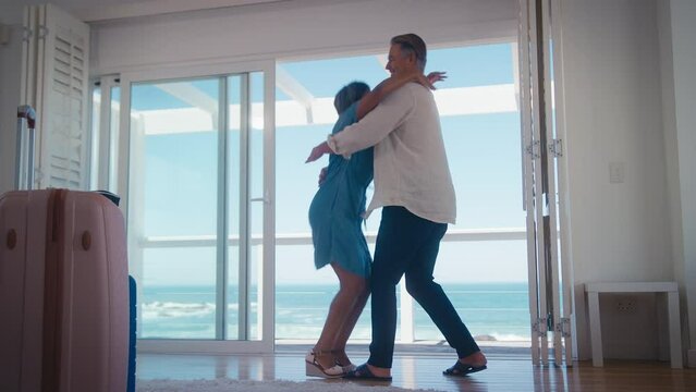 Low angle shot of mature couple with luggage arriving for holiday in beach front property overlooking ocean hugging together - shot in slow motion