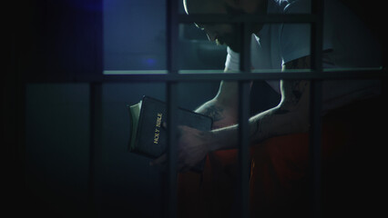 Male prisoner in orange uniform sits on bed in prison cell, reads Bible. Illegally convicted man serves imprisonment term in jail. View through metal bars. Detention center or correctional facility.