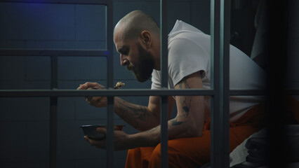 Male prisoner in orange uniform sits on bed in prison cell, eats disgusting prison food from iron...
