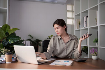Young Asian business woman sits on the phone in an online business meeting using a laptop in a modern home office decorated with shady green plants.