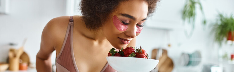 young african american woman in lingerie smelling fresh strawberries in kitchen, banner - obrazy, fototapety, plakaty