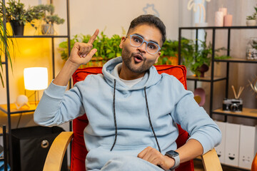 Naklejka premium Eureka. Thoughtful clever inspired Indian man make gesture raises finger came up with creative plan feels excited with good idea inspiration motivation question at home apartment. Guy sits on chair