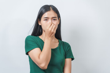 Young Asian frustrated woman with green t-shirt pinches her nose with disgust on her face, reacting to a bad smell in isolation over background.