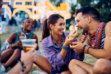 Happy couple having fun while sharing hamburger during open air music festival.