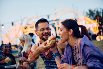 Happy couple sharing hamburger while attending open air music concert in summer.