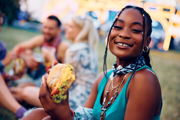 Happy black woman eating burger during open air music festival and looking at camera.