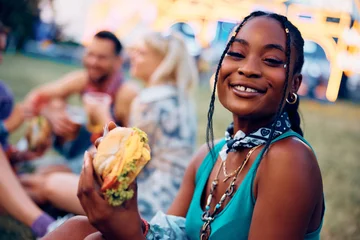 Fototapeten Happy black woman eating burger during open air music festival and looking at camera. © Drazen