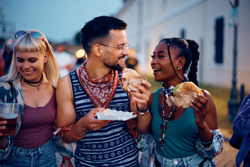 Multiracial group of happy friends eating hamburgers and talking during summer music festival.