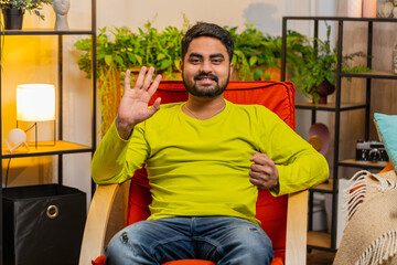 Hello. Indian Hindu man smiling friendly at camera and waving hands gesturing hello hi, greeting or goodbye welcoming with hospitable expression at home. Arabian guy sitting on chair in living room