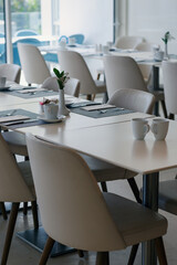 White tables with striped placemats and white cups are paired with chairs. Small vases holding green plants adorn the tables. Large windows allow natural light to illuminate the space. Modern interior