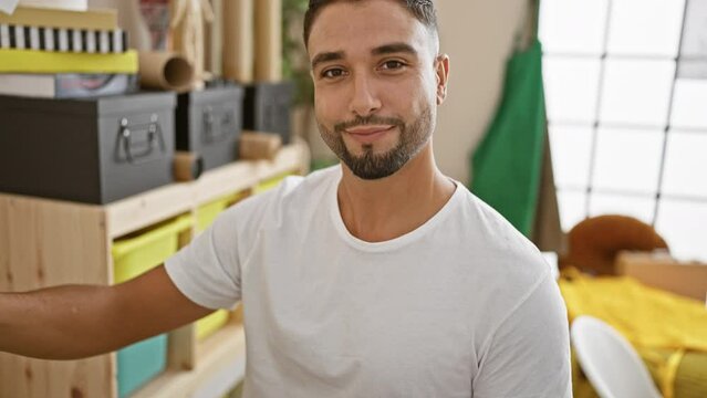 A bearded young man holding a paint palette smiles confidently in a bright creative studio.