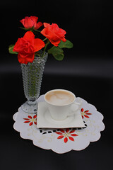 An image of a white cup of coffee on a small plate standing on a white tablecloth, a vase with red flowers, and a dark background.