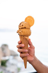 Hand holding yellow ice cream in wafer cone with cookie on the sea background. Eating ice cream in the street
