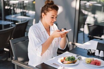 A woman uses a phone and eats lunch or breakfast outdoors in a cafe. Woman eating healthy food on a...