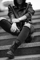Fototapeta premium A beautiful girl with a stylish bob haircut is walking around the city, sitting on the stairs. The woman is wearing a black leather, sweater, torn jeans and boots