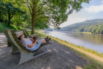 Woman with her dachshund taking a break, lying on large bench, banks of the Prum river at Stausee...