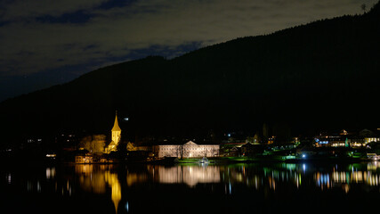 Noc. Oświetlony kościół z wieżą. Odbicia w ciemnej toni jeziora. Ciemny zarys góry. Alpy.