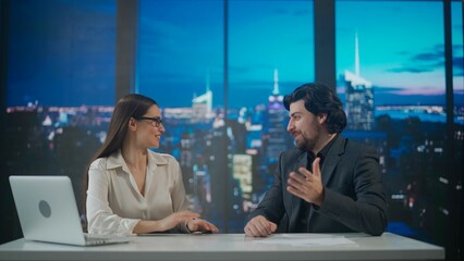 Male and female presenters in the studio. Man and woman news hosts sit at desk presenting evening events, talking joking on air, virtual graphic background at back. © kinomaster