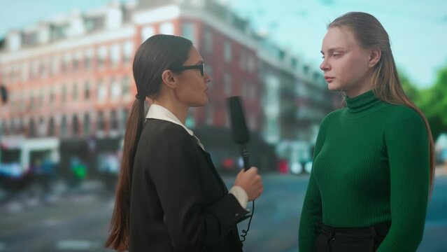 Female presenter reporting on air outside. Woman news host journalist taking interview with young female on the street, city background at the back.