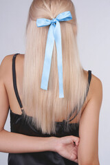 Close-up shot of a young woman with blonde hair and a blue bow hair tie. Portrait of a girl in a black dress and blue bow with long hair. A woman standing with her back on a gray background.