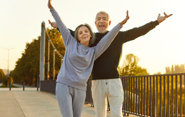 Senior couple having fun during outdoor fitness workout on summer morning. Happy old man and woman in sportswear standing on bridge, feeling full of energy, raising hands up and smiling. Sport concept