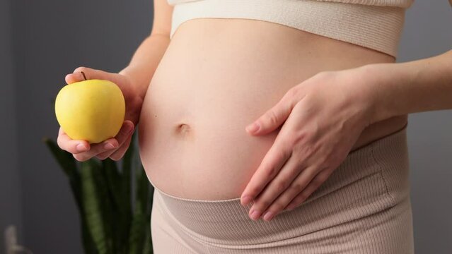 Closeup of belly of pregnant woman holding an apple in her hand enjoying healthy organic eating during pregnancy touching her tummy with love while standing in living room