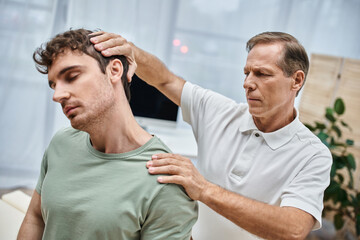appealing mature doctor in uniform massaging his handsome patient during rehabilitation in hospital