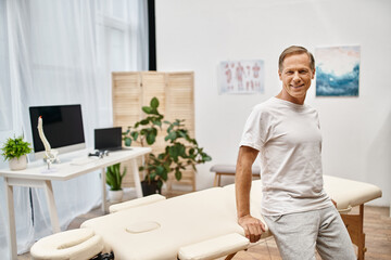 handsome cheerful mature patient in casual attire posing in hospital ward and smiling at camera