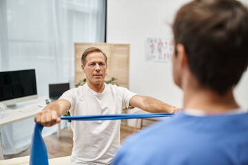 Fototapeta premium focus on joyous mature man in cozy attire using resistance band in front of his blurred doctor