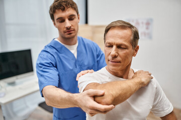 good looking patient in casual attire stretching his muscles with help of his handsome doctor