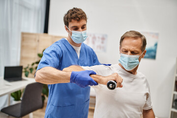 Fototapeta na wymiar hardworking doctor with mask and gloves helping his patient to use dumbbells during appointment