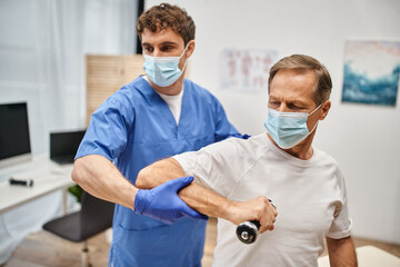 dedicated doctor with mask and gloves helping his patient to use dumbbells on rehabilitation