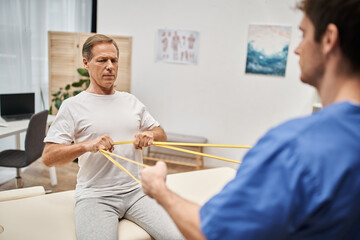 focus on mature patient training with resistance band with his blurred doctor, rehabilitation