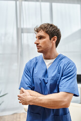 good looking doctor in blue robe posing with arms crossed on chest and looking away in hospital