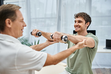 Obraz premium handsome blurred doctor helping cheerful patient in casual attire to recover muscles with dumbbells