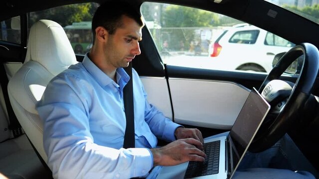Handsome entrepreneur working on notebook while riding an autonomous self driving electric car at urban road. Male businessman typing text on laptop during riding on electrical vehicle with autopilot