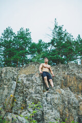 Naklejka na ściany i meble Young muscular man on the background of rocks. Athletic pumped-up man with a bare torso poses against the background of a forest rock.