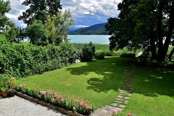 Blick auf den türkisfarbenen Wörthersee in Kärnten mit Blumen, grünem Rasen und Schilfgürtel im Vordergrund