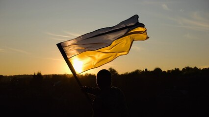 Young man in military uniform waving flag of Ukraine against beautiful sunset at background. Male ukrainian army soldier lifted national banner at countryside. Victory against russian aggression