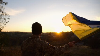 Young man in military uniform waving flag of Ukraine against beautiful sunset at background. Male ukrainian army soldier lifted national banner at countryside. Victory against russian aggression - 759084275